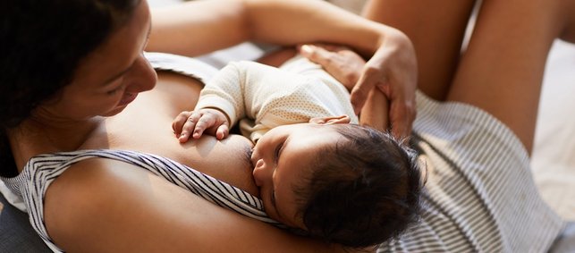 Frau in gestreiftem Sommerpyjama stillt Baby im Liegen