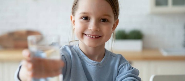 Ein kleines Mädchen mit hellblauem Pullover streckt der Kamera lächelnd ein Glas Wasser entgegen.