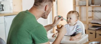 Mann fuettert Baby im Hochstuhl mit Brei aus einem Glaeschen.