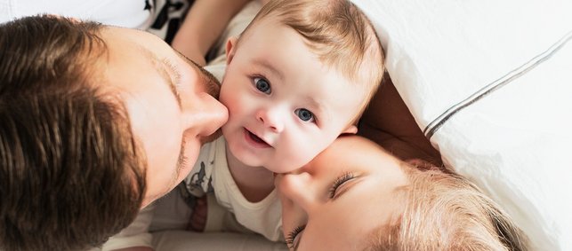 Zufriedenes Baby schaut in die Kamera und wird von Mutter und Vater auf die Wange geküsst