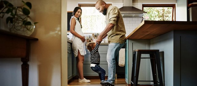 Vater tollt mit Kleinkind in Küche herum und schwangere Frau schaut lächelnd zu