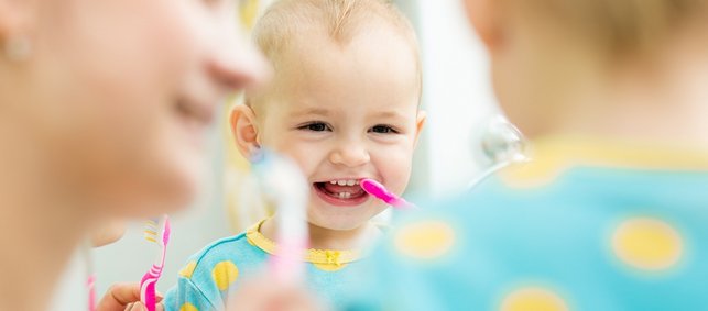 Baby mit ersten Milchzähnen lacht beim Zähneputzen mit Mutter 