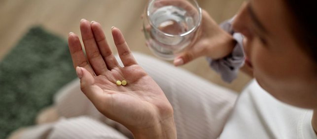 Eine Frau hat Tabletten in der einen Handfläche liegen, in der anderen Hand ein Glas Wasser.