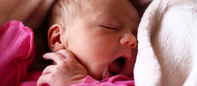 Portrait eines hungrigen Babys in eine weiche Decke gekuschelt, mit geöffnetem Mund und Hand am Gesicht. 