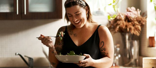 Frau steht in der Küche und isst lachend einen Salat