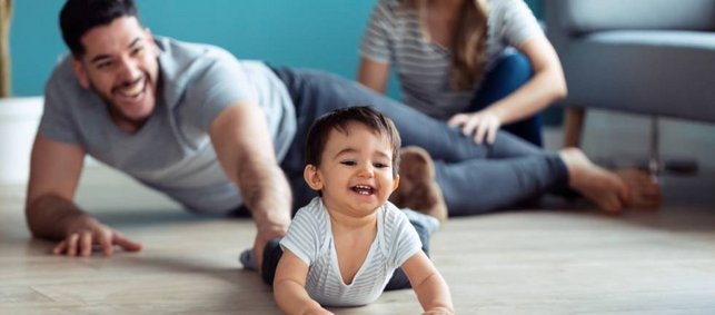 Ein dunkelhaariger kleiner Junge krabbelt lachend über den Fußboden, sein Vater liegt schräg hinter ihm und lacht begeistert, die Mutter sitzt hinter diesem.