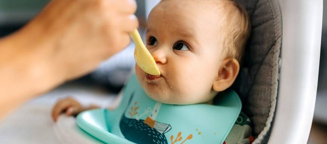 Ein etwa ein halbes jahr altes Baby sitzt in einem Hochstuhl und wir mit einem Löffel Brei gefüttert.