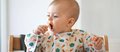 Baby in Hochstuhl isst mit den Händen ein gegartes Stück Gemüse, wahrscheinlich Pastinake. 