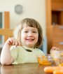 Ein etwa dreijähriges Mädchen sitzt am Tisch, hält einen Löffel in der Hand und isst Karottensalat. Im Vordergrund liegen Karotten auf dem Tisch.