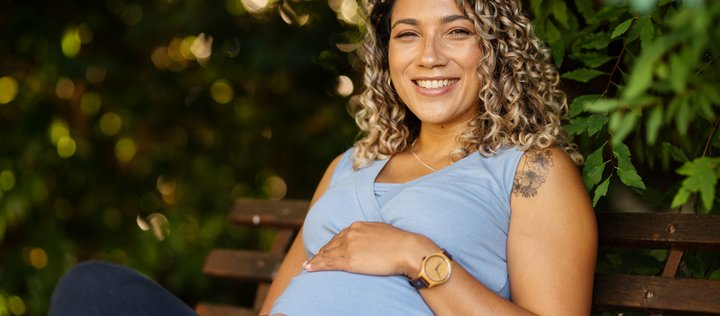 Schwangere lockige Frau auf Parkbank lächelt und halt die Hände auf dem Bauch