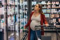 Eine Schwangere steht suchenden Blickes in einem Supermarkt vor dem Kühlregal, die Hand auf einem Eikaufswagen abgelegt. 