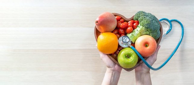 Obst und Gemüse liegen in einer herzförmigen Schale, die jemand in der Hand hält. Darum ist ein Stethoskop drapiert.