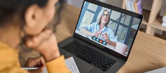 Dunkelhaarige Frau schaut auf einen Laptop-Bildschirm, worin eine ältere Frau zu sehen ist, die etwas vorträgt..