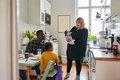 In einer Küche sitzen ein Mann und ein Kind am Tisch, eine Frau mit Baby im Arm steht ihnen zugewandt daneben.