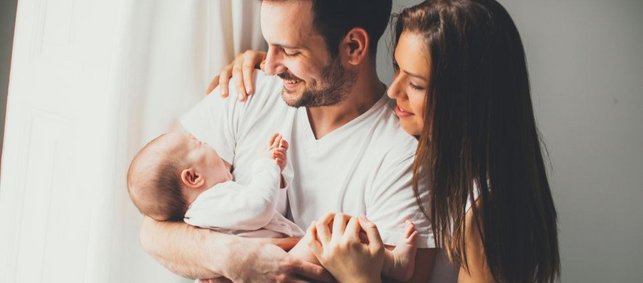 Vater hält Neugeborenes im Arm, die Mutter steht umamend hinter dem Mann. Beide schauen ihr Neugeborenes verliebt an. 