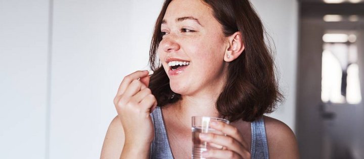 Junge Frau mit braunen Haaren und Sommersprossen hält ein Glas Wasser in der einen hand und führt mit der anderen eine Tablette zu ihrem Mund.