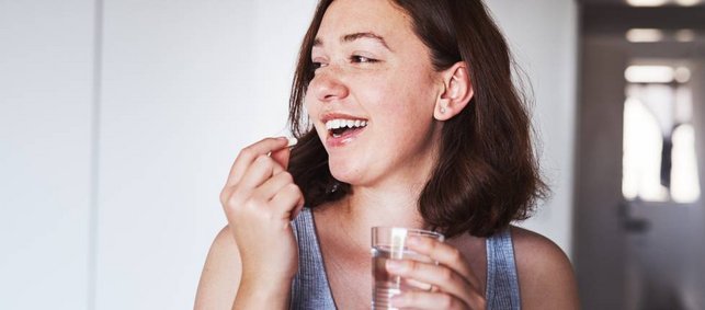 Junge Frau mit braunen Haaren und Sommersprossen hält ein Glas Wasser in der einen hand und führt mit der anderen eine Tablette zu ihrem Mund.
