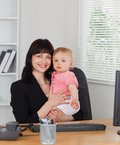 Eine junge Frau hält am Schreibtisch sitzend ein Baby im Arm. Beide lächeln in die Kamera.