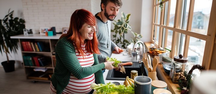 Ein schwangeres Paar steht gut gelaunt in der Küche und bereitet Gemüse zum Kochen vor.