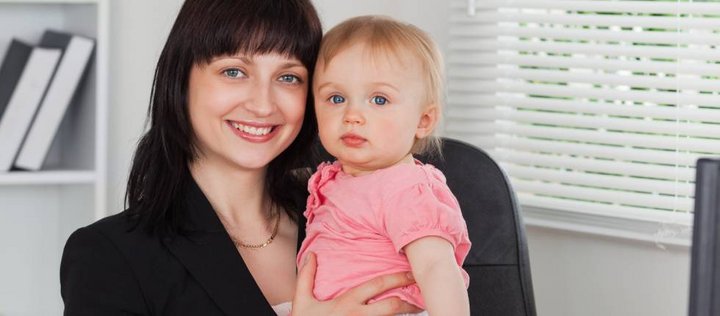 Eine junge Frau sitzt mit einem Baby im Arm in einem Büro am Schreibtisch. Beide lächeln in die Kamera.