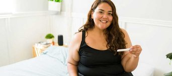 Eine lächelnde Frau mit Übergewicht sitzt auf dem Bett und hält einen positiven Schwangerschaftstest in der Hand. 