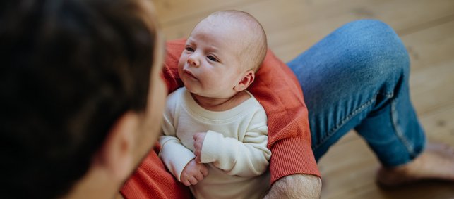 Mann sitzt auf dem Boden in der Wohung und hält neugeborenes Baby auf dem Schoss, das ihn ansieht