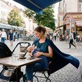 Eine Frau stillt ein Baby in einer Fußgängerzone. Sie trägt einen dunkelblauen Umhang.