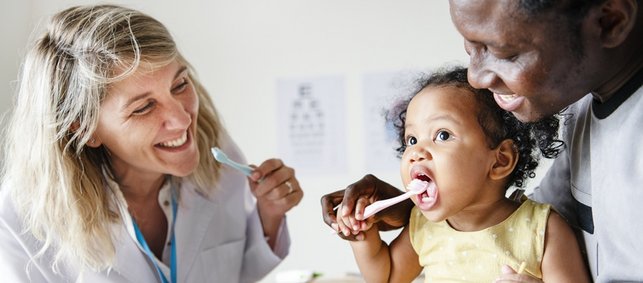 Zahnärztin zeigt Kleinkind mit Vater Zähneputzen