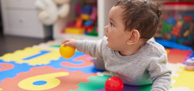Baby liegt in Bauchlage auf einer Zahlen-Matte, einen kleinen Ball vor sich und einen in der Hand.