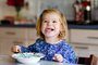 Ein etwa zweijähriges Mädchen sitzt am Tisch und isst Milchbrei aus einer Schüssel. Sie grinst schelmisch.