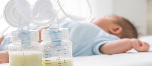 Zwei Milchpumpen im Vordergrund, zur Hälfte gefüllt mit Muttermilch. Im Hintergrund liegt ein Baby.