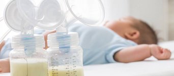 Zwei Milchpumpen im Vordergrund, zur Hälfte gefüllt mit Muttermilch. Im Hintergrund liegt ein Baby.